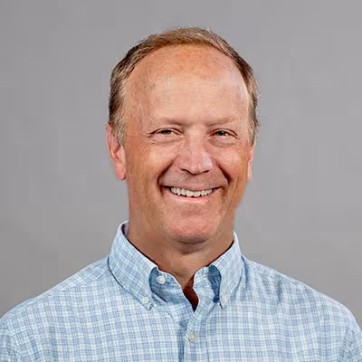 J. Andrew Berglund smiles in a light blue checked button-up shirt in front of a gray backdrop