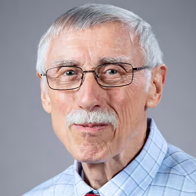 Ray Bromley smiles in glasses and a blue plaid shirt and red tie against a gray background.