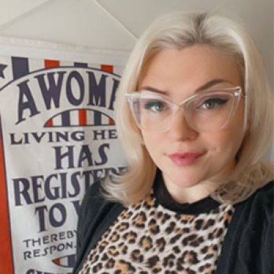 Samantha Hall-Saladino looks into the camera wearing clear-framed glasses, a leopard-print top and black cardigan. Behind her, a red white and blue sign is slightly cut-off, but reads,"A Woma.../Living he.../Has /Register.../To V.../Thereby.../Respons..."