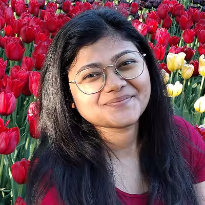Saumya Gupta smiles in front of a bed of tulips while wearing glasses and a red short sleeved shirt