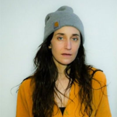 Tara Autovino wears a yellow button-down shirt and a gray beanie as she looks into the camera against a plain background.