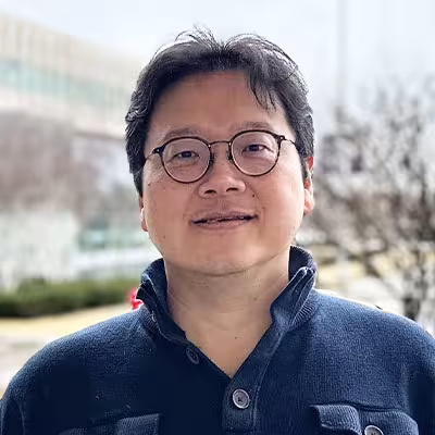 Tse-Chuan Yang stands on UAlbany campus wearing glasses and a navy pullover.