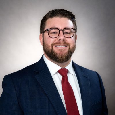 Smiling portrait of Michael Colello in a dark suit with a white shirt and red tie. 