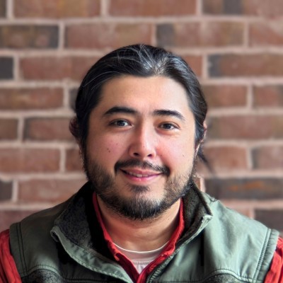 Arthur wearing a red shirt with a green vest sitting in front of a brick wall