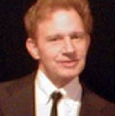 Portrait of Paul wearing a dark suit and tie with a white shirt and black background. 