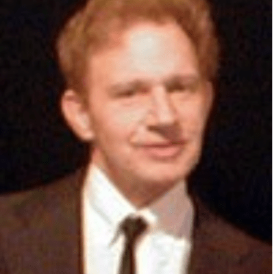 Portrait of Paul wearing a dark suit and tie with a white shirt and black background. 