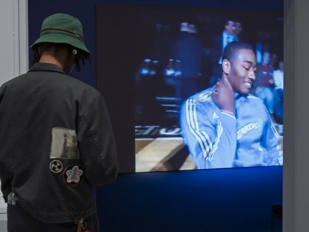 Person in a jacket and bucket hat in foreground. Projection of a basketball player dancing in the background. 