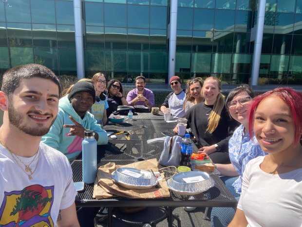 Ten members of STEM NOW gather for a lunch meet up outside in the Life Sciences courtyard on a sunny day. They are seated around a picnic table scattered with assorted food containers and water bottles. All students are smiling.