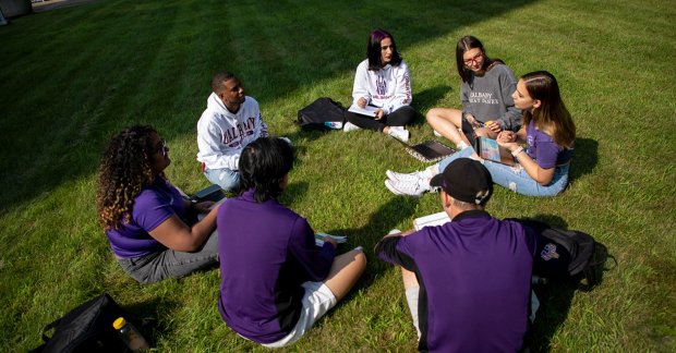 Seven students wearing UAlbany t-shirts and sweatshirts sit in a circle on a grassy part of campus.
