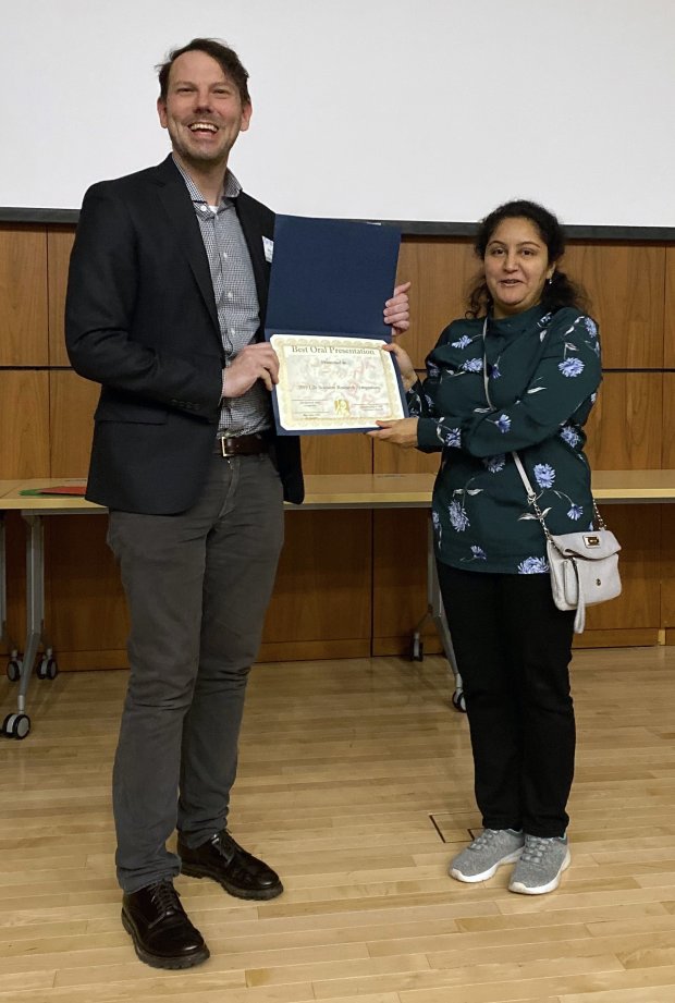 A woman in a green top and black pants stands to the right and accepts an award certificate from a man in a suit jacket.