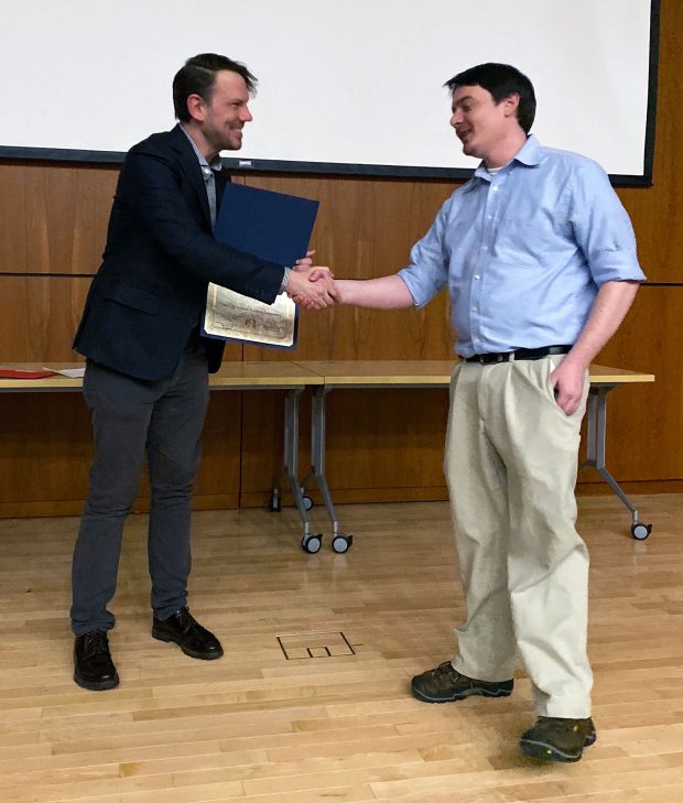 A man in a blue shirt and khakis shakes hands and accepts an award certificate from a man in a suit jacket.
