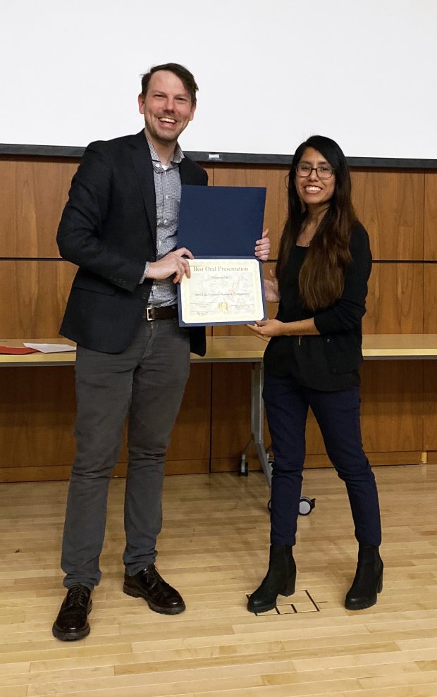 A woman in black stands to the right and accepts an award certificate from a man in a suit jacket.