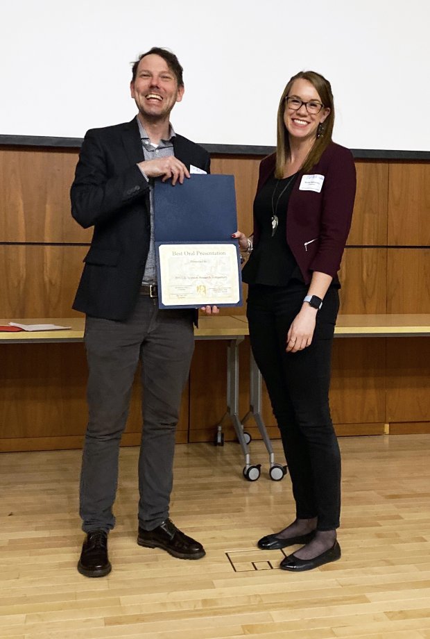 A woman in black with a maroon cardigan stands to the right and accepts an award certificate from a man in a suit jacket.