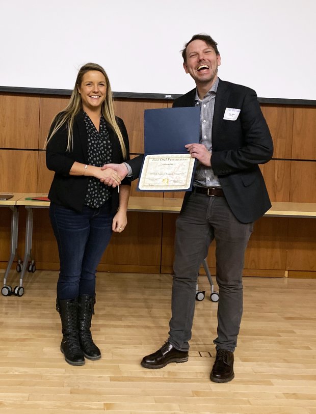 A woman in a patterned top, black cardigan and jeans shakes hands with accepts an award certificate from a man in a suit jacket.