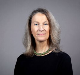 Margi Sheehy in black top and green neckace with shoulder length hair, gray background