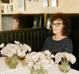 Lynn Tillman sits in a booth at a restaurant in a gray patterned blouse and glasses. Three vases with while flowers sit on a table before her.
