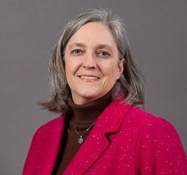 Billie Franchini smiles wearing a burgundy turtleneck and red blazer against a gray backdrop