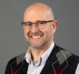 Justin Albohn smiles in glasses, a checked button up and argyle sweater vest against a gray backdrop