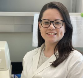 Amy Mascorro stands in a lab. smiling in glasses and a white lab coat