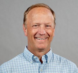 J. Andrew Berglund smiles in a light blue checked button-up shirt in front of a gray backdrop