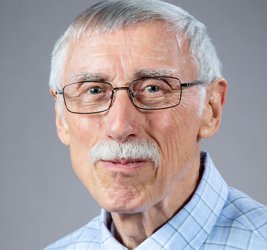 Ray Bromley smiles in glasses and a blue plaid shirt and red tie against a gray background.