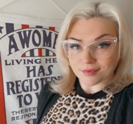 Samantha Hall-Saladino looks into the camera wearing clear-framed glasses, a leopard-print top and black cardigan. Behind her, a red white and blue sign is slightly cut-off, but reads,"A Woma.../Living he.../Has /Register.../To V.../Thereby.../Respons..."