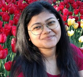Saumya Gupta smiles in front of a bed of tulips while wearing glasses and a red short sleeved shirt
