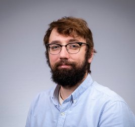 Jon smiles at the camera, standing against a grey background.
