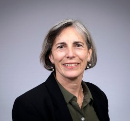 Janice Pata is standing against a grey wall and wears a black suit jacket.