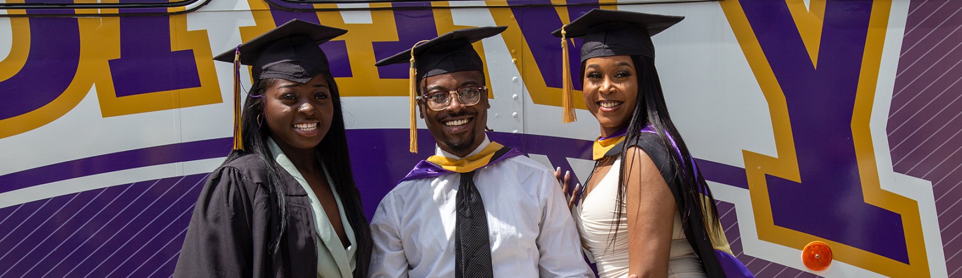 Three graduating students standing in front of a UAlbany bus.