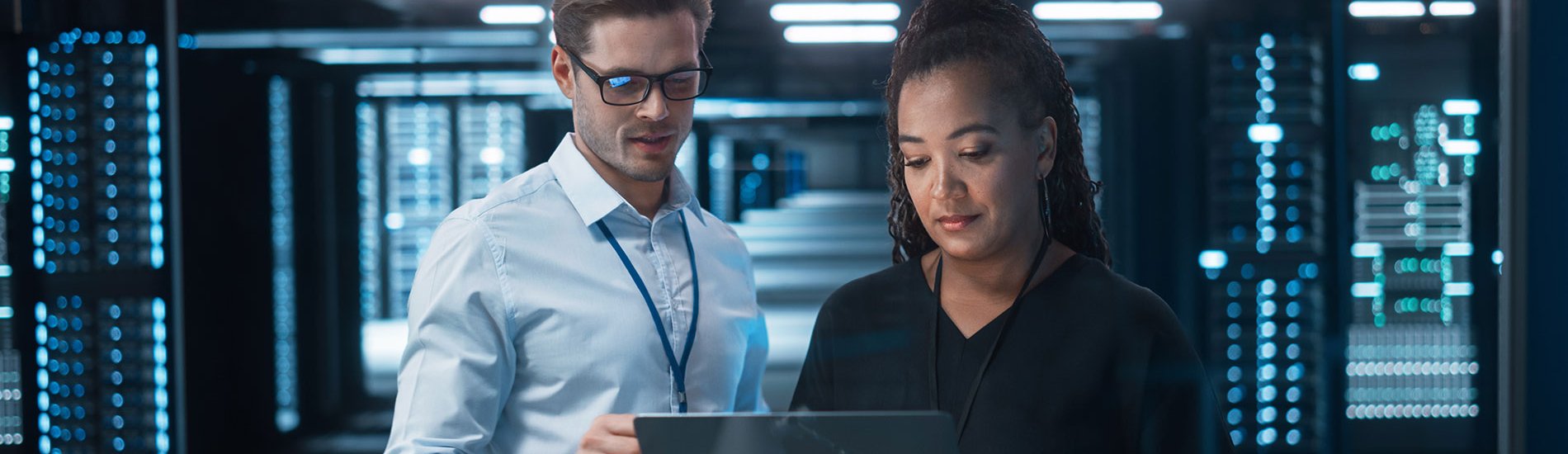 Two cybersecurity professionals standing in a server room consult a laptop.