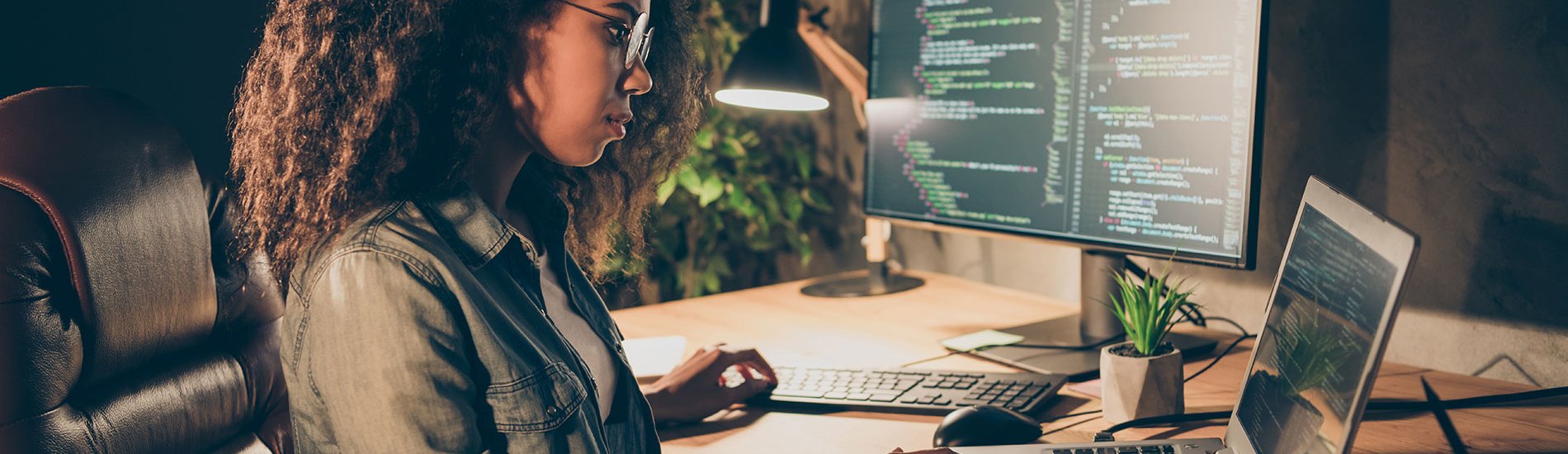 A cybersecurity professional works on a laptop alongside a monitor displaying code.