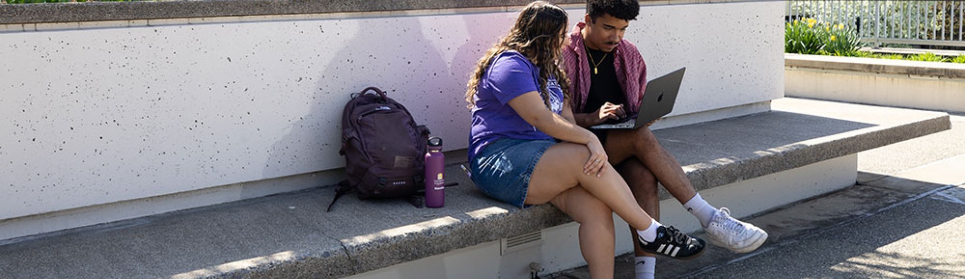 Two students sitting together on a stone bench looking at a laptop.