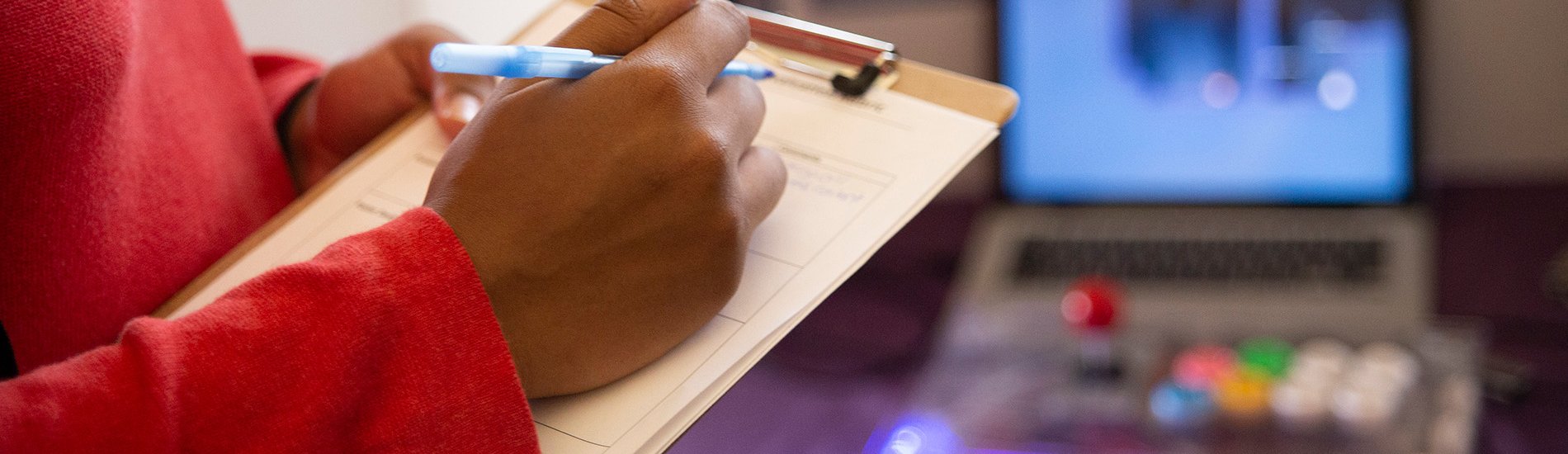 A hand poised to write on a clipboard, with a computer system visible in the background