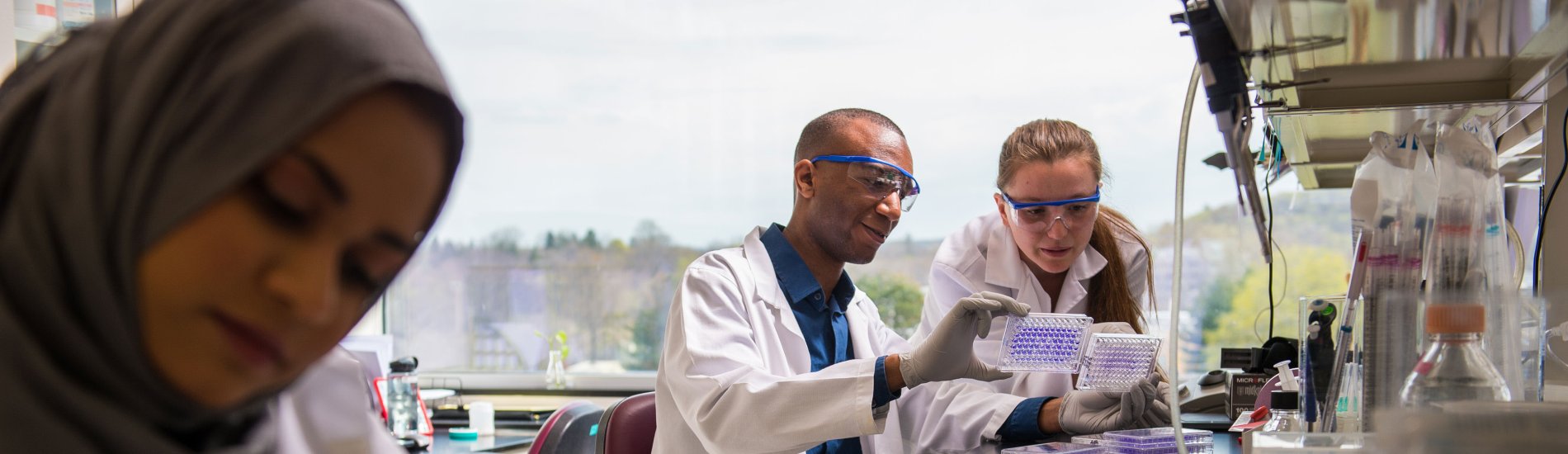Two researchers in white lab coats and protective glasses examine a specimen in the background, while a third researcher writes in a notebook in the foreground.