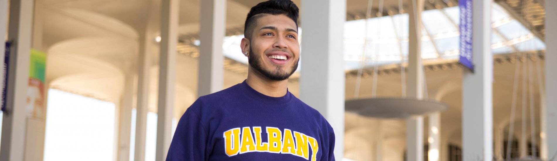 A student wearing a purple UAlbany sweatshirt smiles and poses for a photo on campus, with the Podium arches visible behind him