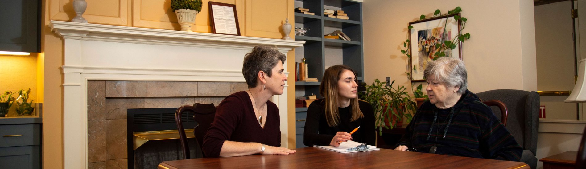 Three adults of varying age sit at a table inside a sitting room and speak.