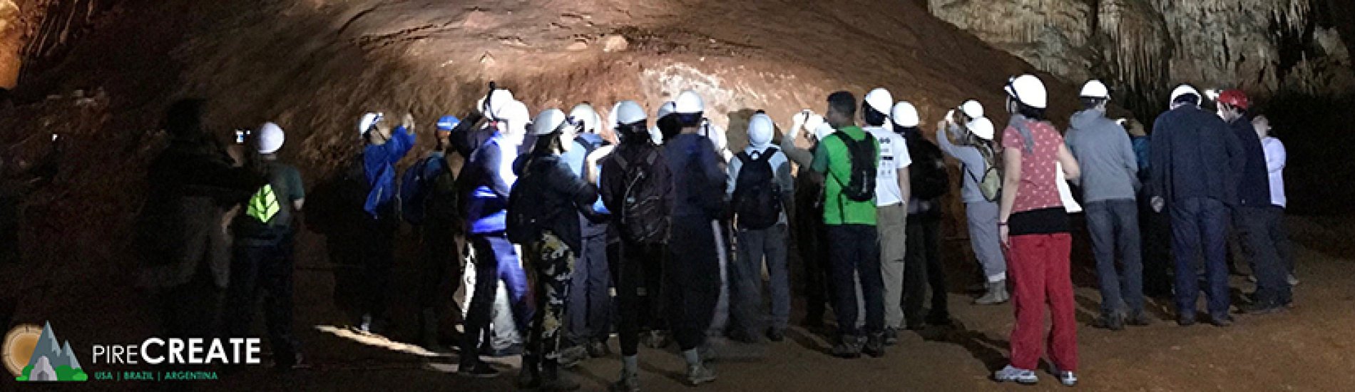 A group of students gather in a cavern while wearing head lamps and looking up at stalactites hanging from the ceiling. 