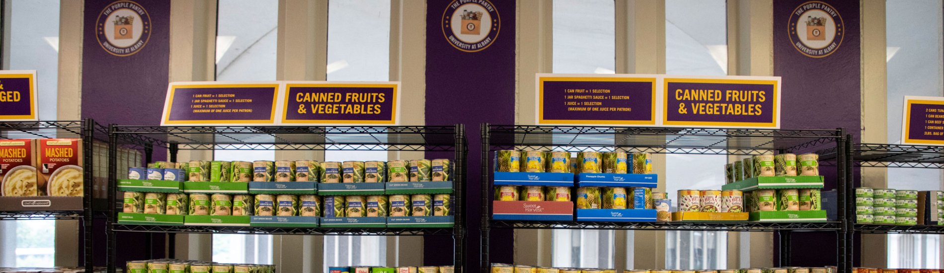 Shelves stocked with canned fruit and vegetables inside the Purple Pantry. Signs show the Purple Pantry logo and instructions for community members.