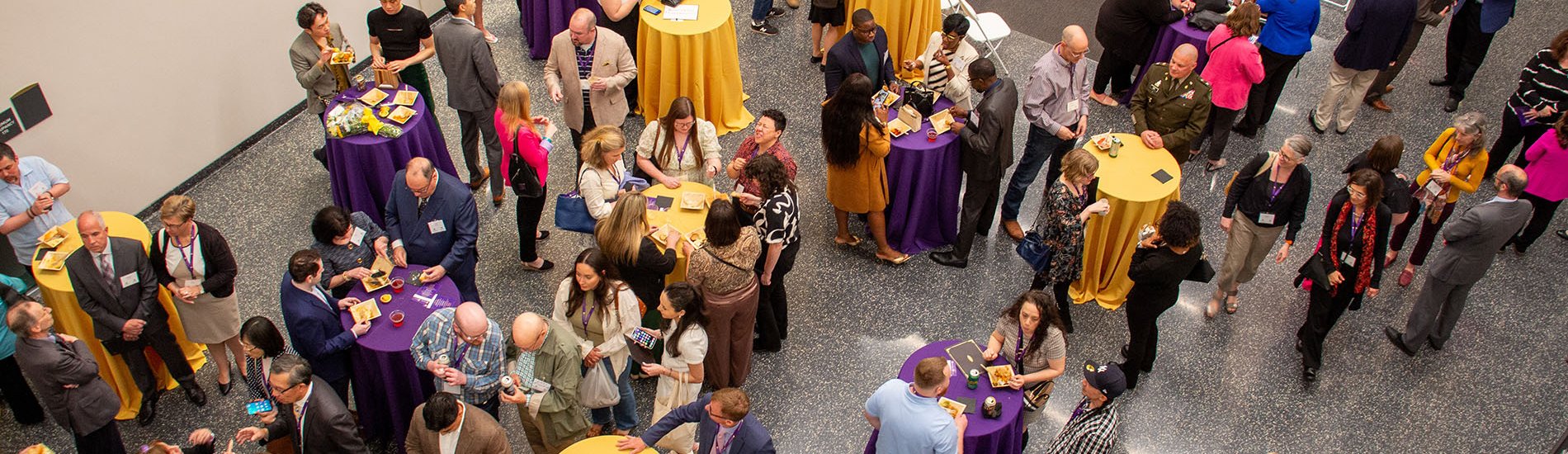 An image looking down at individuals standing around purple and yellow high tables at the Rockefeller Colleges' SCJ Awards Ceremony and Reception.