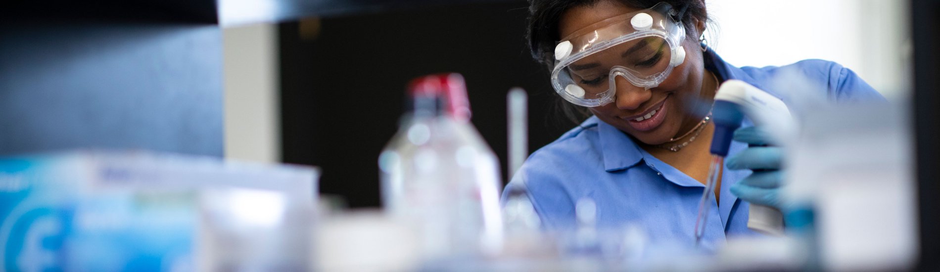 A smiling researcher in safety googles, gloves and jacket uses a device to prepare samples for research.