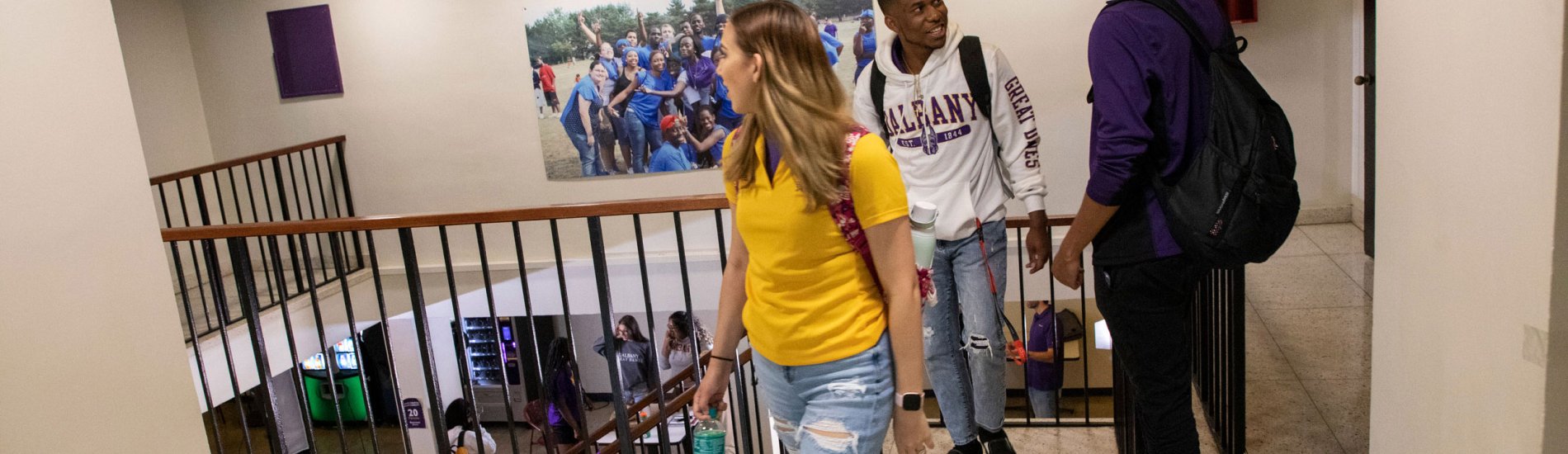 Three students walk by a stairwell inside a residence hall. Vending machines are visible downstairs.