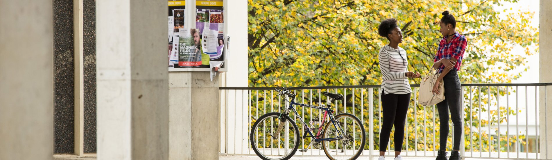 Two students stand outside a residential quad talking and smiling on a spring day. There is a bicycle and a bulletin board in the background.