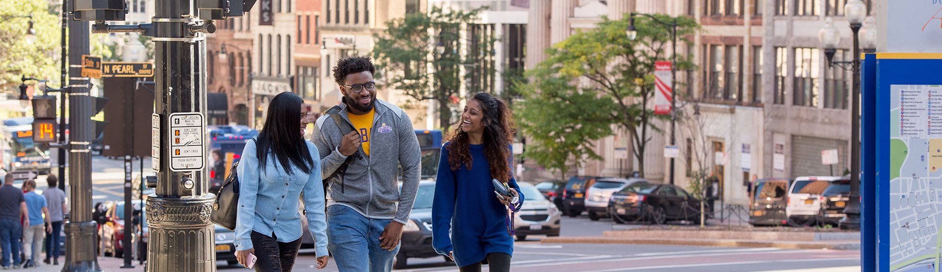Three students smile as they walk along a downtown Albany city street.