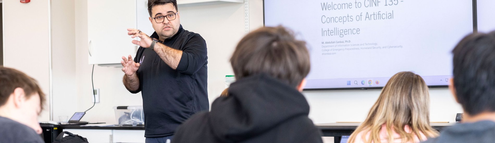 Dr. M. Abdullah Canbaz gestures as he teaches a classroom full of students. The screen behind him shows the course name, CINF 135: Concepts of Artificial Intelligence.