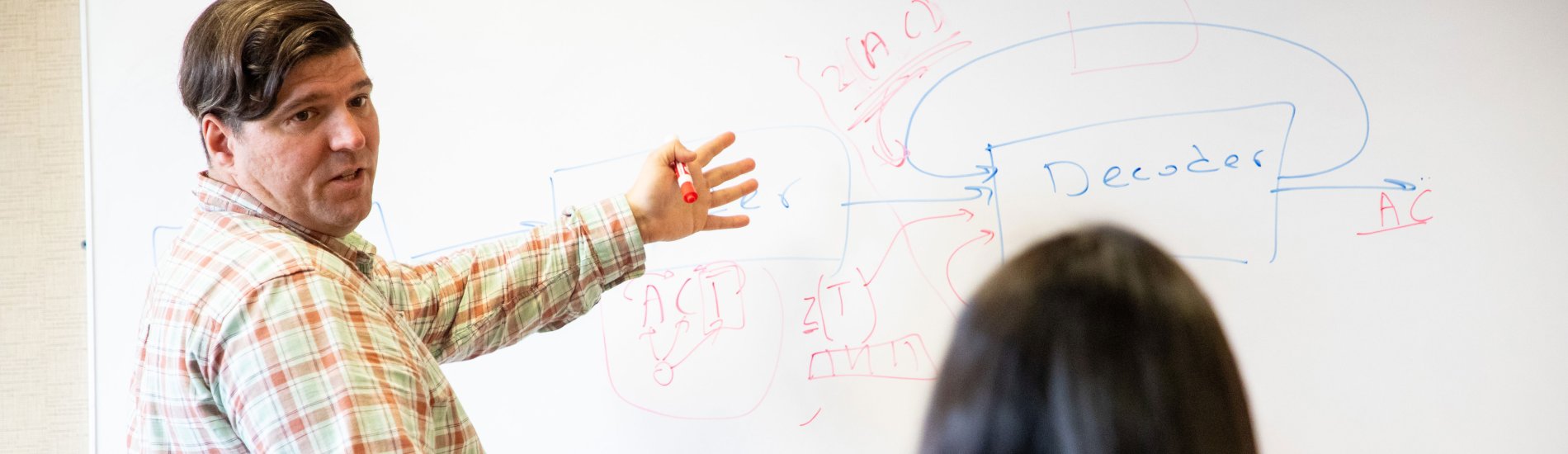 A UAlbany professor gestures toward a white board, where he's drawn a sketch labeled "Decoder." A seated student is visible in the foreground.