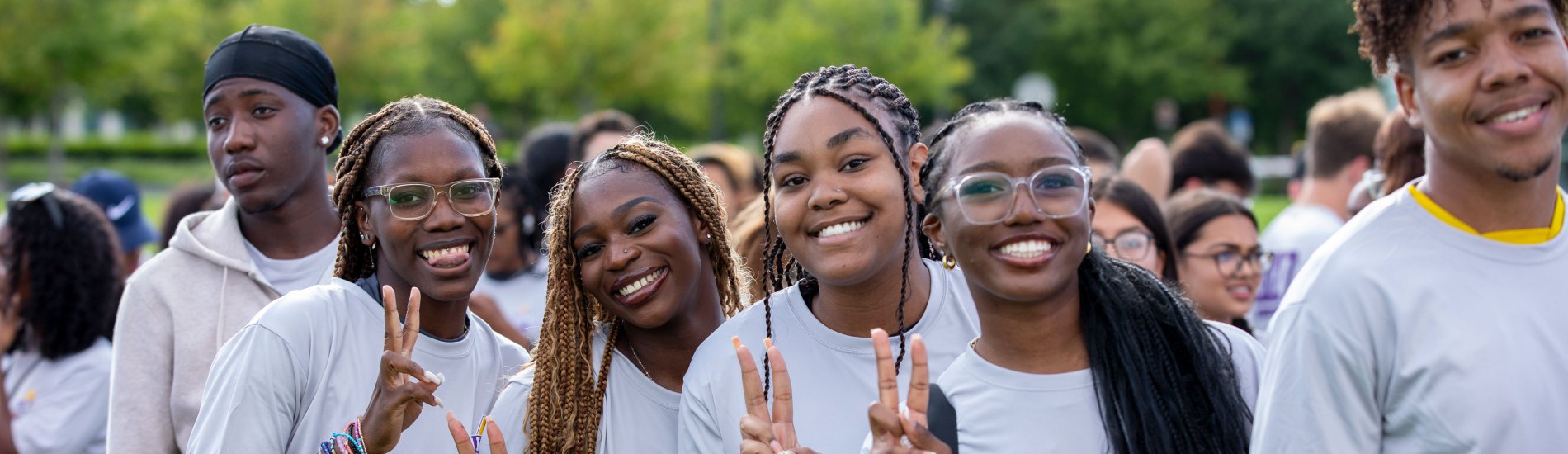 Six Black students wearing matching light gray t-shirts smile and pose for a photo. Four of the students hold up peace signs.