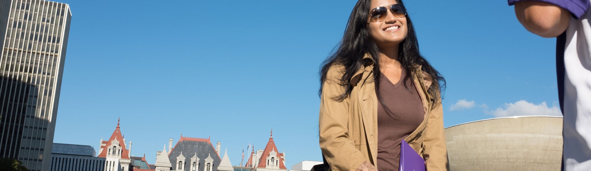 A student wearing sunglasses and a light coat smiles as she stands in front of the New York State Capitol building.