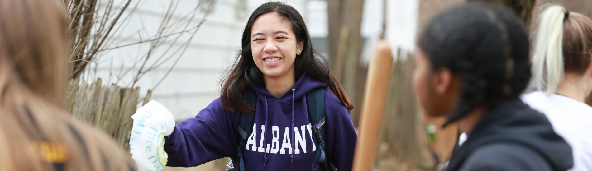 A smiling student wearing a purple UAlbany sweatshirt reaches out to throw away a piece of trash in Albany's Pine Hills neighborhood.