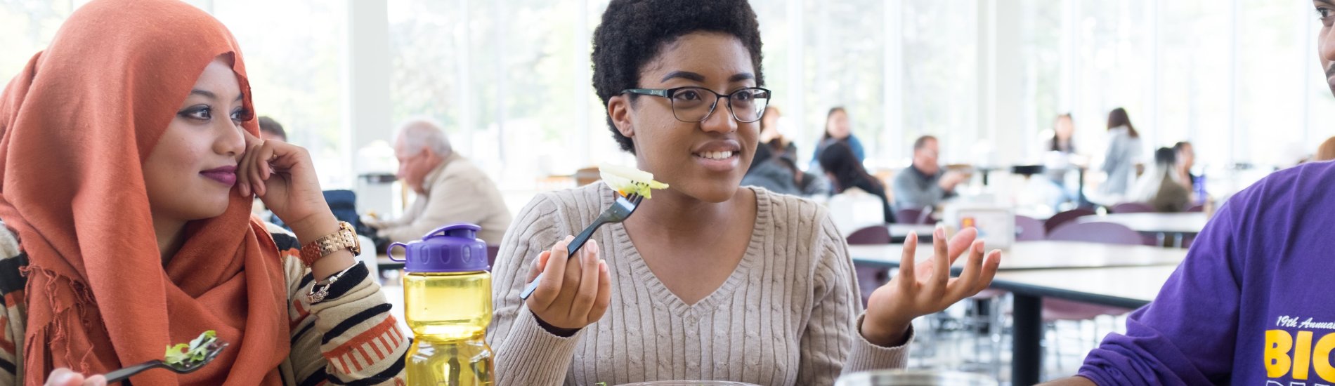 Three students gesture, smile and talk while sitting at a table eating salads.